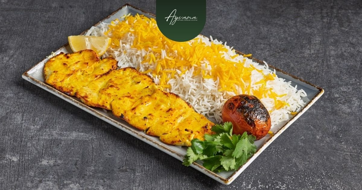 Plate of saffron rice with grilled Joojeh Kabab, roasted tomato, lemon, and herbs on dark restaurant table backdrop