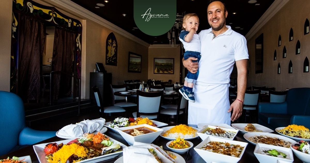 Smiling chef holding his young son in Ayvana Persian Restaurant with a table full of colorful Persian dishes