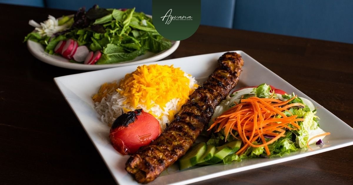 Grilled Persian kabab with saffron rice, charred tomato, fresh salad, and shredded carrots elegantly plated on a white square dish at Ayvana.