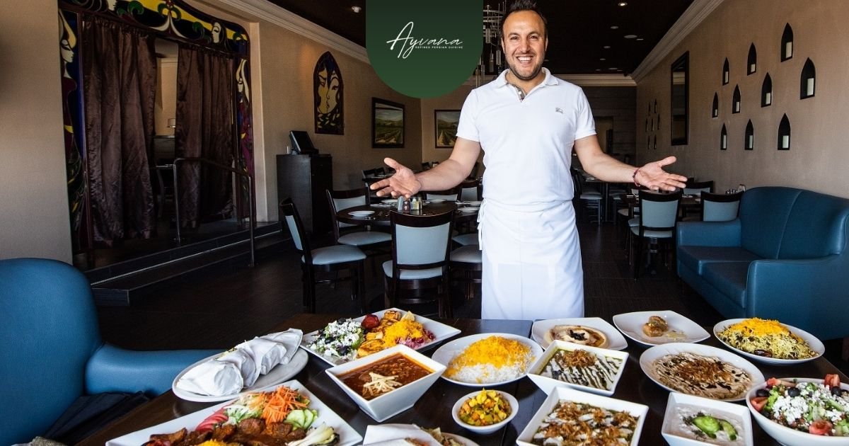 Smiling chef in white apron welcomes guests at Ayvana Persian Restaurant in Tarzana, proudly displaying a table filled with assorted Persian kebabs and traditional dishes.